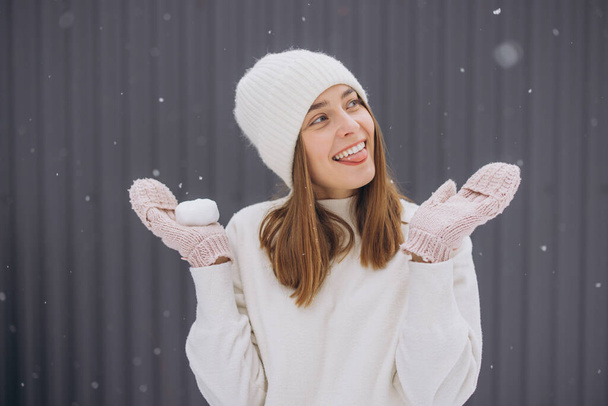 Счастливая женщина в вязаных варежках пытается поймать снежинки языком на сером фоне зимой - Фото, изображение