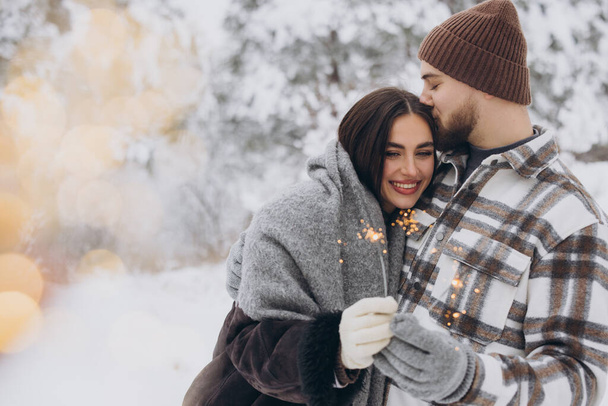 Pareja romántica sonriente en sombreros de punto posando con chispas en el bosque de invierno, sosteniendo luces de bengala en las manos, celebrando vacaciones de Navidad juntos, Copiar el espacio - Foto, Imagen