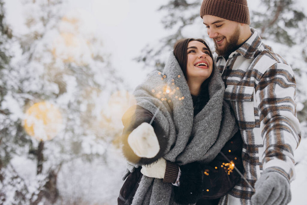Usmívající se romantický pár v pletených klobouky pózování s jiskry v zimním lese, držení bengálských světel v rukou, slaví vánoční svátky spolu, kopírovat prostor - Fotografie, Obrázek
