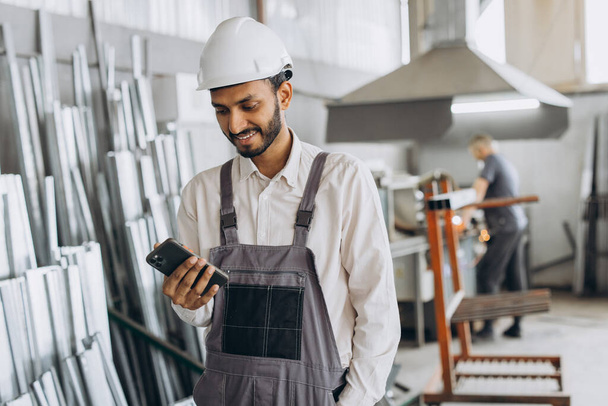 Szczęśliwy brodaty indyjski pracownik fabryki mężczyzn w specjalnym mundurze i biały twardy kapelusz trzymając telefon i mając dialog w produkcji okien i drzwi metalowo-plastikowych - Zdjęcie, obraz