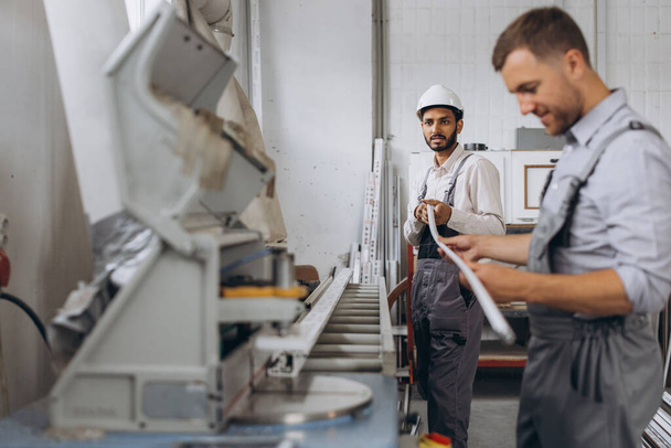 Un equipo internacional, dos trabajadores trabajan en una máquina en una fábrica de ventanas y puertas de PVC - Foto, Imagen