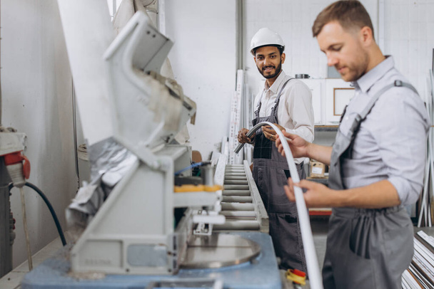 Een internationaal team, twee arbeiders werken bij een machine in een fabriek van PVC ramen en deuren - Foto, afbeelding