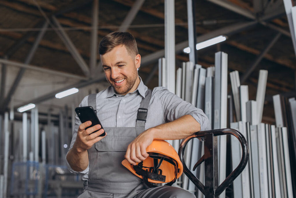 Portrait d'un travailleur heureux dans un casque orange et salopettes tenant un camion hydraulique et parlant au téléphone sur un fond d'usine et cadres en aluminium. - Photo, image