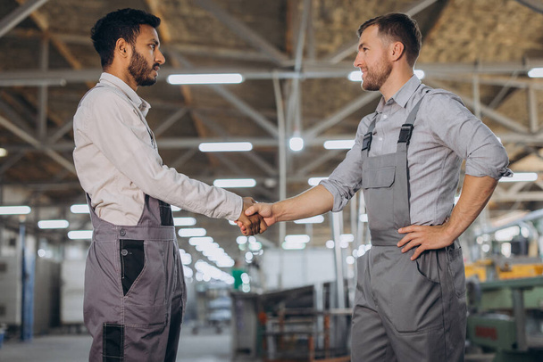 Due lavoratori internazionali felici in tuta intera stringono la mano su uno sfondo di fabbrica - Foto, immagini