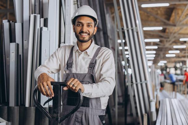 Portret szczęśliwego hinduskiego pracownika w białym, twardym kapeluszu i kombinezonie trzymającego ciężarówkę hydrauliczną na tle fabryki i aluminiowych ram. - Zdjęcie, obraz