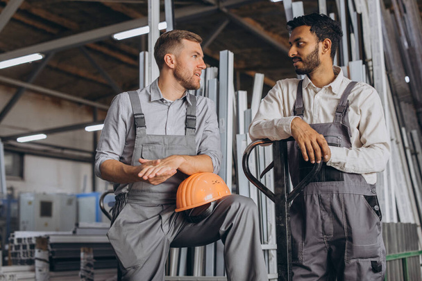 Portret van twee internationale arbeiders die een helm dragen die pauze neemt van hun werk en met elkaar praat op een fabrieksachtergrond met kopieerruimte - Foto, afbeelding
