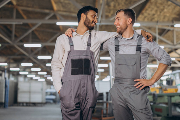 Two happy international workers in overalls hug each other against a factory background - Photo, Image