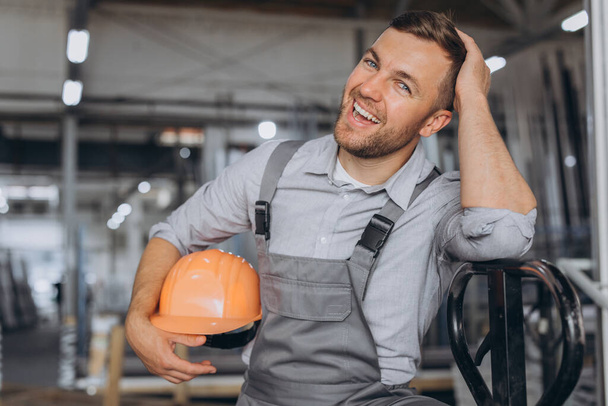 Retrato de un trabajador feliz con un sombrero duro naranja y un mono que sostiene un camión hidráulico sobre un fondo de una fábrica y marcos de aluminio. - Foto, imagen