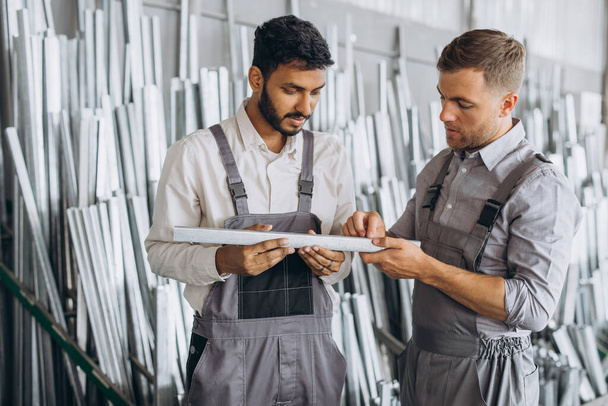 Dwóch międzynarodowych pracowników w mundurach omawia proces pracy w fabryce okien metalowo-plastikowych - Zdjęcie, obraz
