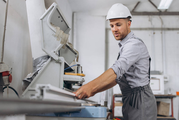 Een mannelijke werknemer in een speciaal uniform en een witte helm werkt op een CNC-machine voor de productie van PVC ramen en deuren in de fabriek. - Foto, afbeelding