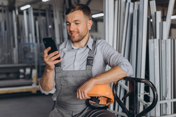 Portrait of a happy worker in a orange helmet and overalls holding a hydraulic truck and talking on the phone against a background of a factory and aluminum frames. - Photo, Image