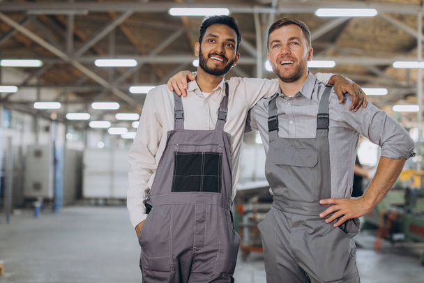 Dwóch szczęśliwych międzynarodowych pracowników w kombinezonach przytula się do siebie na tle fabryki. - Zdjęcie, obraz