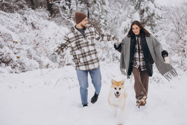 Happy young couple with akita dog in forest on winter and snowly day - Photo, Image