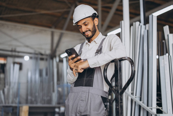 Portrait of a happy Hindu worker in a white helmet and overalls holding a hydraulic truck and talking on the phone against a background of a factory and aluminum frames. - Photo, Image