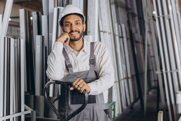 Portret szczęśliwego hinduskiego pracownika w białym, twardym kapeluszu i kombinezonie trzymającego ciężarówkę hydrauliczną na tle fabryki i aluminiowych ram. - Zdjęcie, obraz