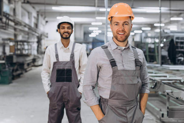 Dos trabajadores masculinos internacionales en overoles en el fondo de la producción de ventanas y puertas de PVC - Foto, imagen