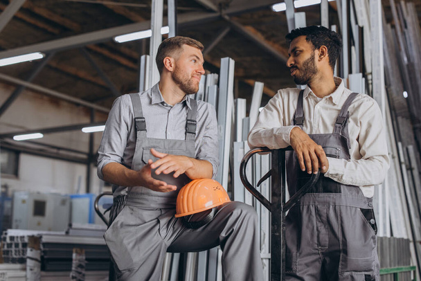 Προσωπογραφία δύο διεθνών εργαζομένων με σκληρά καπέλα που κάνουν διάλειμμα από την εργασία και αναπαύονται μιλώντας ο ένας στον άλλο σε ένα εργοστάσιο φόντο με αντίγραφο χώρο - Φωτογραφία, εικόνα