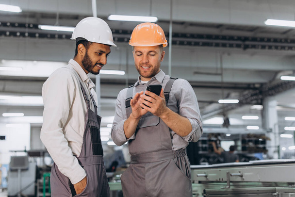 Due lavoratori maschi internazionali in tuta intera guardando il telefono sullo sfondo della produzione di finestre e porte in PVC in alluminio - Foto, immagini
