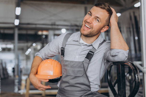 Portrait d'un travailleur heureux portant un casque orange et une salopette tenant un camion hydraulique sur fond d'usine et de cadres en aluminium. - Photo, image