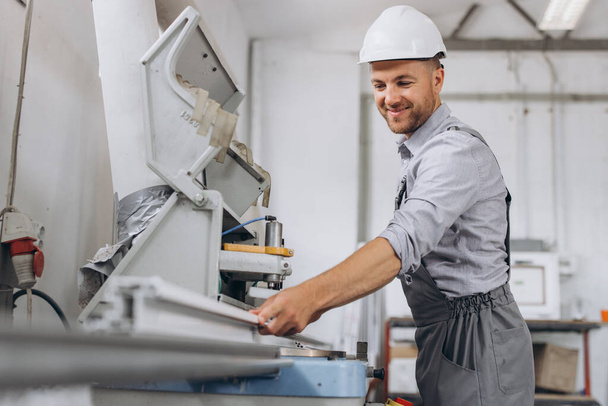 Un trabajador masculino con un uniforme especial y un casco blanco trabaja en una máquina CNC para la producción de ventanas y puertas de PVC en la fábrica. - Foto, imagen