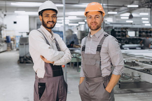 Dos trabajadores masculinos internacionales en overoles en el fondo de la producción de ventanas y puertas de PVC - Foto, imagen
