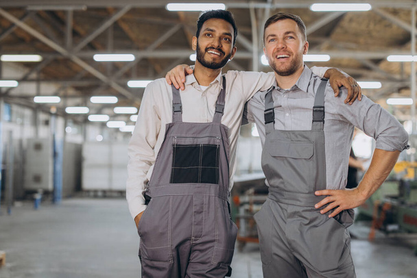 Twee blije internationale arbeiders in overalls knuffelen elkaar tegen een fabrieksachtergrond - Foto, afbeelding