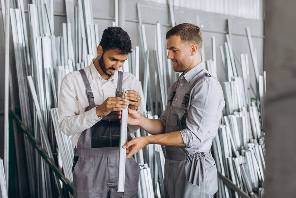 Deux travailleurs masculins internationaux en uniforme discutent du processus de travail dans une usine de fenêtres métal-plastique - Photo, image