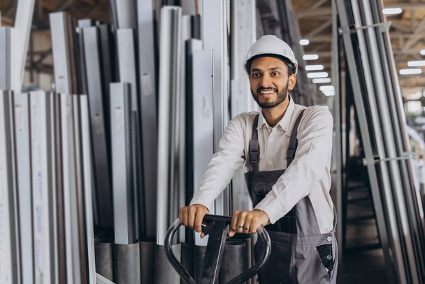 Portrait d'un heureux travailleur hindou portant un casque blanc et une salopette tenant un camion hydraulique sur fond d'usine et de cadres en aluminium. - Photo, image
