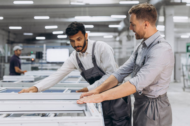 Due lavoratori internazionali maschi in tuta da lavoro lavorano insieme in una macchina per la produzione di finestre e porte in PVC in alluminio - Foto, immagini