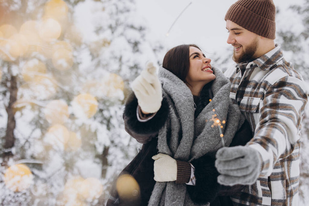 Χαμογελώντας ρομαντικό ζευγάρι με πλεκτά καπέλα ποζάροντας με λαμπάδες στο Χειμερινό Δάσος, κρατώντας φώτα της Βεγγάλης στα χέρια, γιορτάζοντας τις διακοπές των Χριστουγέννων μαζί, αντιγραφή χώρου - Φωτογραφία, εικόνα