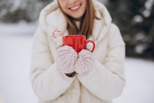 Χαριτωμένη ευτυχισμένη γυναίκα σε πλεκτά γάντια κρατώντας ζεστό καφέ με καραμέλα μπαστούνια στο πάρκο το χειμώνα - Φωτογραφία, εικόνα