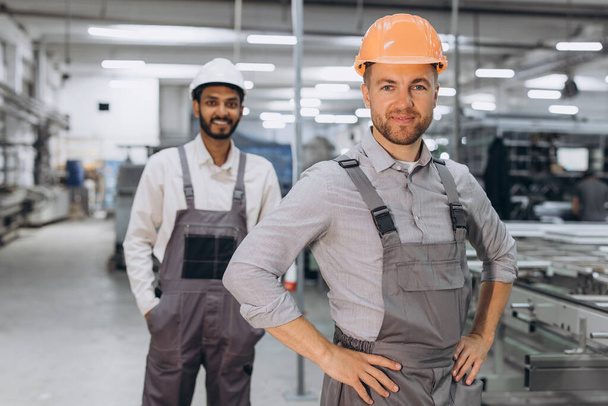 Due lavoratori internazionali maschili in tuta sullo sfondo della produzione di finestre e porte in PVC - Foto, immagini