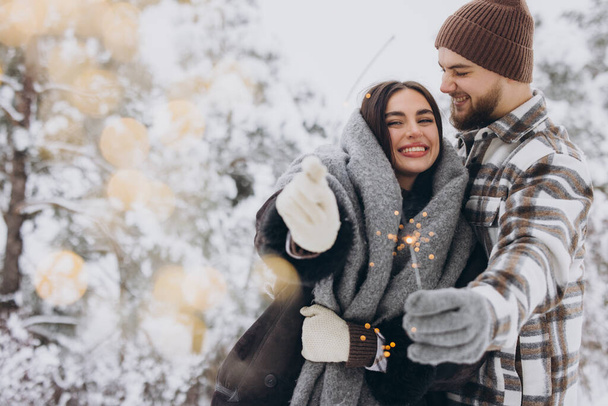 Sorridente coppia romantica in maglia cappelli in posa con scintille a foresta invernale, Tenere le luci del Bengala nelle mani, Celebrare le vacanze di Natale insieme, Copia spazio - Foto, immagini