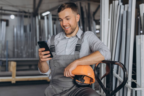 Porträt eines glücklichen Arbeiters mit orangefarbenem Helm und Overall, der einen hydraulischen LKW hält und vor dem Hintergrund einer Fabrik und Aluminiumrahmen telefoniert. - Foto, Bild