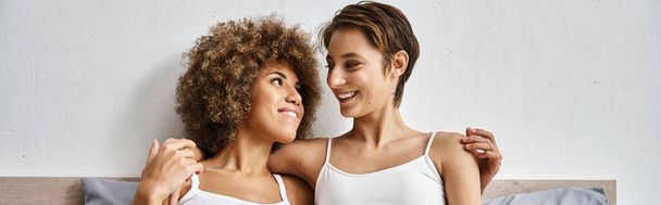 cheerful and multicultural lesbian couple hugging while resting together on bed, horizontal banner - Photo, Image