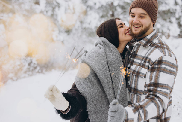 Couple romantique souriant en bonnets tricotés posant avec des étincelles à la forêt d'hiver, tenant les lumières du Bengale dans les mains, célébrant ensemble les vacances de Noël, Copier l'espace - Photo, image