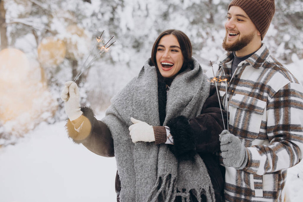 Usmívající se romantický pár v pletených klobouky pózování s jiskry v zimním lese, držení bengálských světel v rukou, slaví vánoční svátky spolu, kopírovat prostor - Fotografie, Obrázek