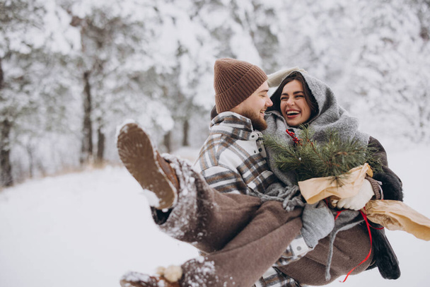 Симпатичная молодая пара, влюбленная в сосновый букет, проводит время в день Святого Валентина в снежном зимнем лесу в горах. Парень держит девушку на руках, улыбаясь друг другу.. - Фото, изображение