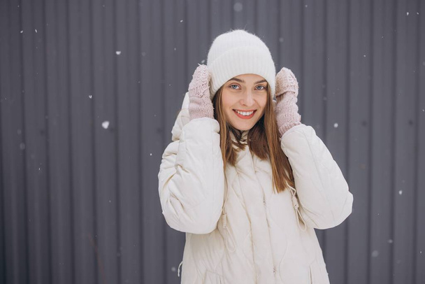 Una mujer feliz en manoplas de punto posa sobre un fondo de una pared gris en invierno en una nevada - Foto, Imagen