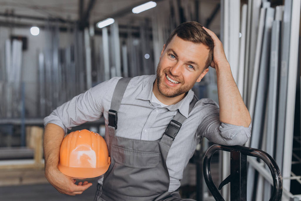 Turuncu şapkalı ve tulumlu mutlu bir işçinin portresi bir fabrika ve alüminyum çerçevelerin arka planında hidrolik bir kamyon tutuyor.. - Fotoğraf, Görsel