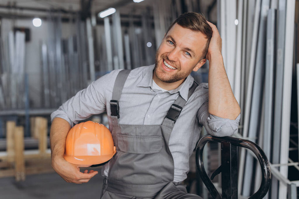 Portret szczęśliwego pracownika w pomarańczowym, twardym kapeluszu i kombinezonie z wózkiem hydraulicznym na tle fabryki i aluminiowych ram. - Zdjęcie, obraz