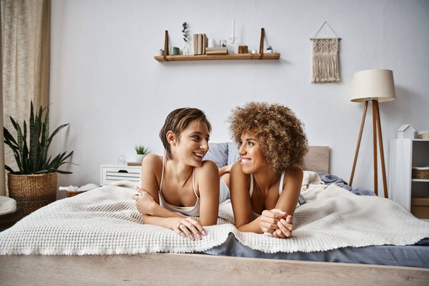 happy lesbian partners looking at each other while lying together on bed at home, cozy moments - Фото, изображение