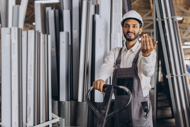 Retrato de un trabajador hindú feliz con un sombrero blanco y un mono que sostiene un camión hidráulico sobre un fondo de una fábrica y marcos de aluminio. - Foto, Imagen
