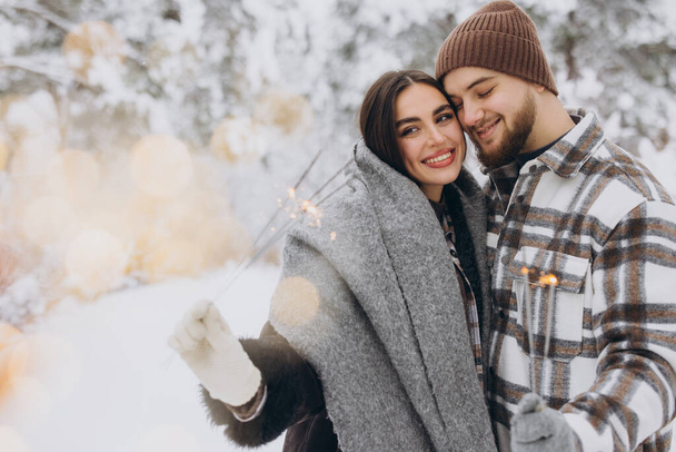 Χαμογελώντας ρομαντικό ζευγάρι με πλεκτά καπέλα ποζάροντας με λαμπάδες στο Χειμερινό Δάσος, κρατώντας φώτα της Βεγγάλης στα χέρια, γιορτάζοντας τις διακοπές των Χριστουγέννων μαζί, αντιγραφή χώρου - Φωτογραφία, εικόνα
