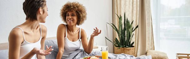 happy multicultural lesbian couple in pajamas gesturing and chatting near breakfast in bed, banner - Photo, Image