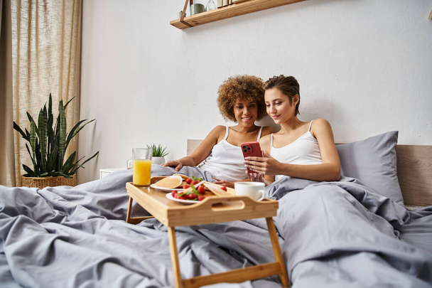 alegre pareja lésbica multicultural en pijama usando smartphone cerca del desayuno en bandeja en la cama - Foto, imagen