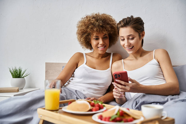 счастливая мультикультурная лесбийская пара в пижаме, используя смартфон рядом с завтраком на подносе в постели - Фото, изображение