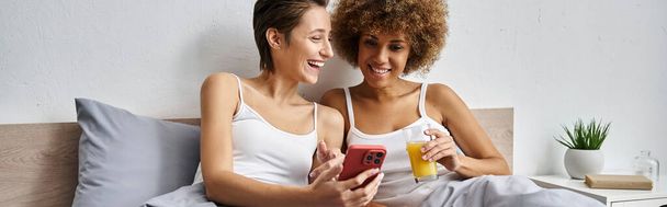 щаслива лесбіянка сміється і використовує смартфон біля афроамериканської дівчини, банер - Фото, зображення