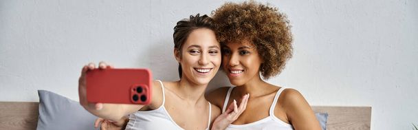 szczęśliwa kobieta biorąc selfie na smartfonie w pobliżu Afryki amerykańskiej dziewczyny w sypialni, baner - Zdjęcie, obraz