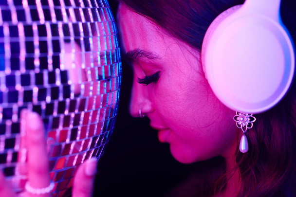 Jonge vrouw luisteren naar muziek in koptelefoon en leunend voorhoofd op discobal - Foto, afbeelding